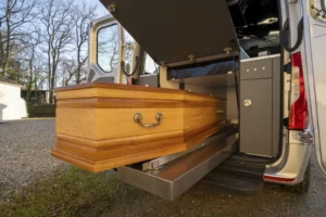 Cercueil en bois clair en cours de chargement sur un plateau coulissant à l'arrière d'un corbillard